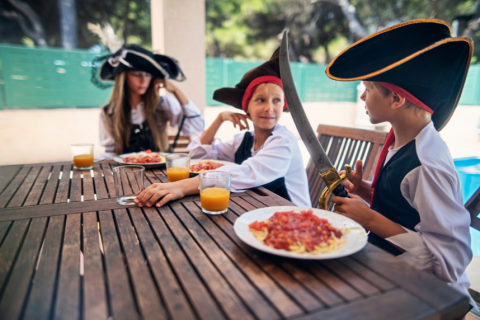 Three children dressed as pirates sitting at a table with spaghetti and orange juice.