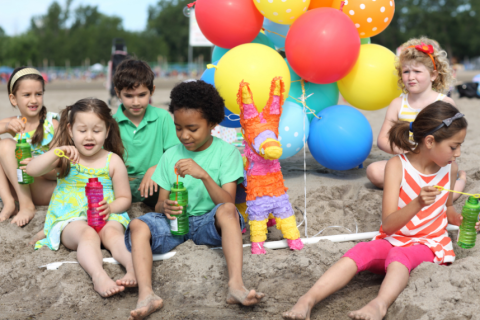 Six children sitting around at the beach blowing bubble wands with balloons and a pinata.