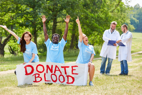 three people kneeling behind a Donate Blood banner. People are smiling with arms raised.