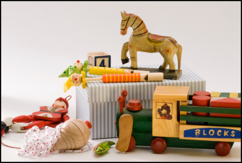 The image shows a group of vintage wooden toys: a carved horse on a striped box, colorful wooden whistles, alphabet blocks, a green and yellow toy train labeled “BLOCKS,” a red pull-toy figure, and a small painted spinning top.