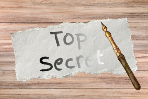 The image shows a torn piece of white textured paper lying on a wooden surface. On the paper, the words “Top Secret” are written in a mix of gray and black lettering with a casual, hand-drawn look fading. To the right side of the paper is a vintage-style fountain pen or dip pen with a metal nib and a wooden handle, positioned diagonally so the nib points upward. The wooden surface beneath has a light brown, rustic appearance with horizontal planks.