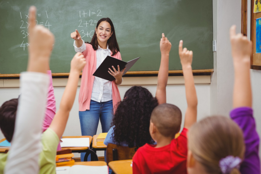 A class of children raising their hands in a classroom and a teacher pointing to a student to call on them to speak.