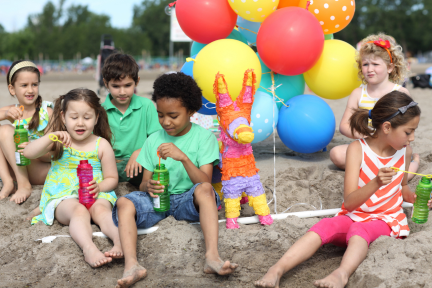 Six children sitting around at the beach blowing bubble wands with balloons and a pinata.