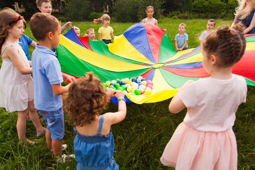 Children standing around, and holding, a rainbow parachute that has multicolored balls on top of it.